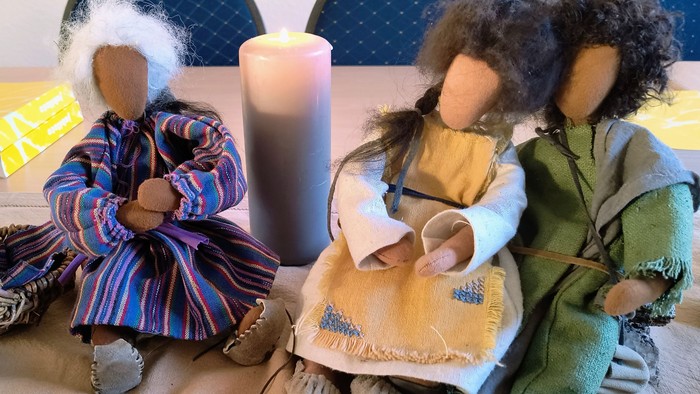 Drei Puppen sitzen auf einem Bett, eine hält ein Baby, eine Kerze steht daneben