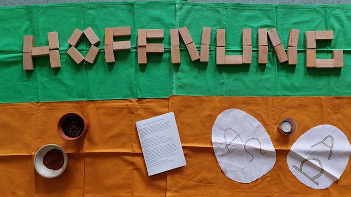 Ein Tisch mit Kaffee, Buch und Bastelmaterialien vor einem grün-orangen Tuch mit der Aufschrift "Hoffnung".