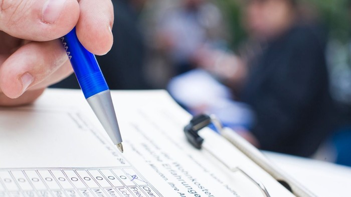 Hand schreibt mit blauem Stift auf Papier, im Hintergrund sind zwei Personen.