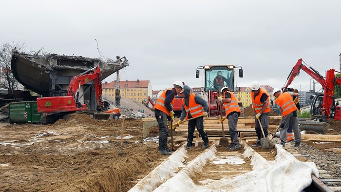 Bauarbeiter in Schutzausrüstung heben mit schwerem Gerät und Werkzeugen in der Nähe einen Graben in städtischer Umgebung aus.
