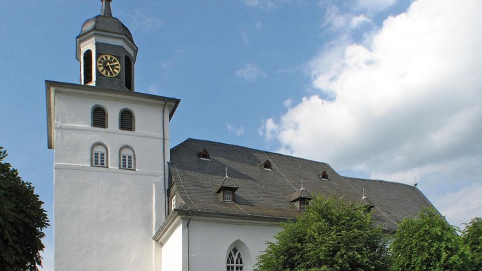 Weiße Kirche mit hohem Turm und Uhr, umgeben von Bäumen