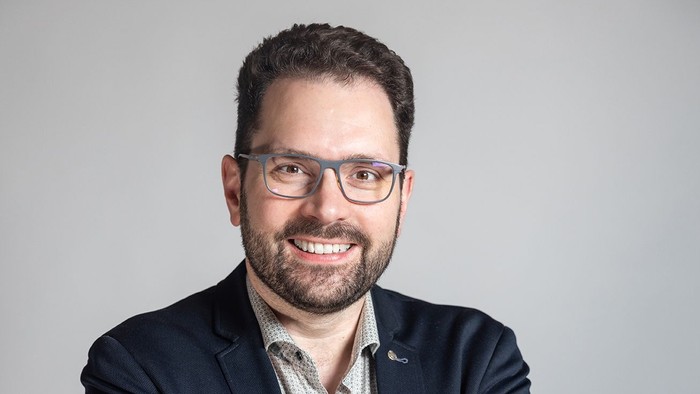 Ein Mann mit Brille und dunklem Jackett lächelt selbstbewusst.