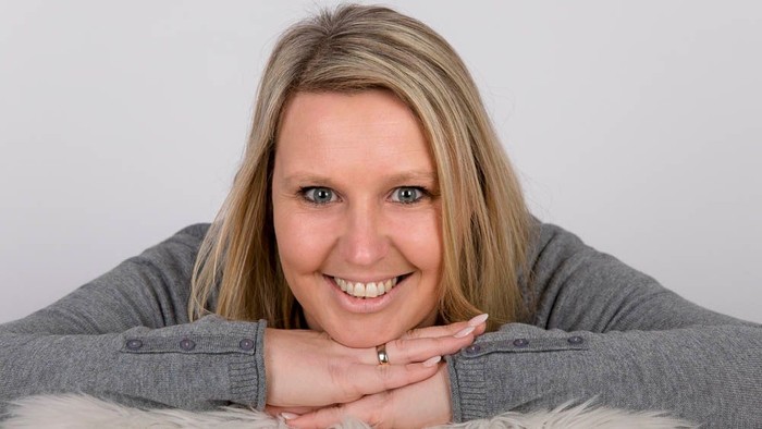 Eine Frau mit blondem Haar lächelt in die Kamera und stützt ihr Kinn auf die Hände.