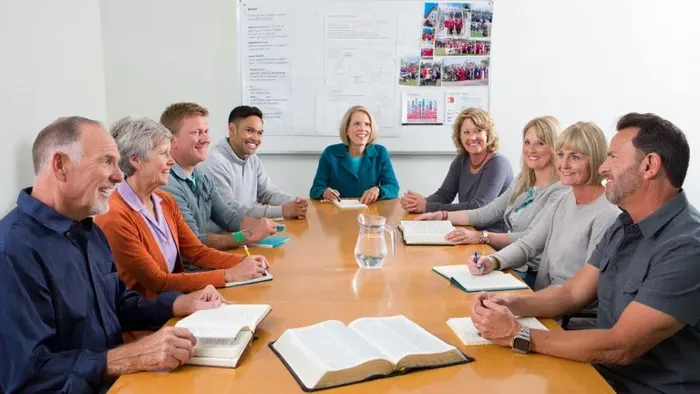 Ein Gemeindekirchenrat bei einer Sitzung in einem schlichten, hellen Raum mit Holztisch. Männer und Frauen mittleren und höheren Alters, diverse Kleidung zwischen Alltags- und Business-Look, diskutieren engagiert. Auf dem Tisch liegen Bibel, Notizbücher, 