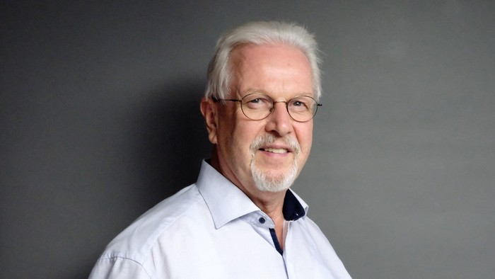 Ein älterer Mann mit Brille und grauem Bart, der ein weißes Hemd trägt.