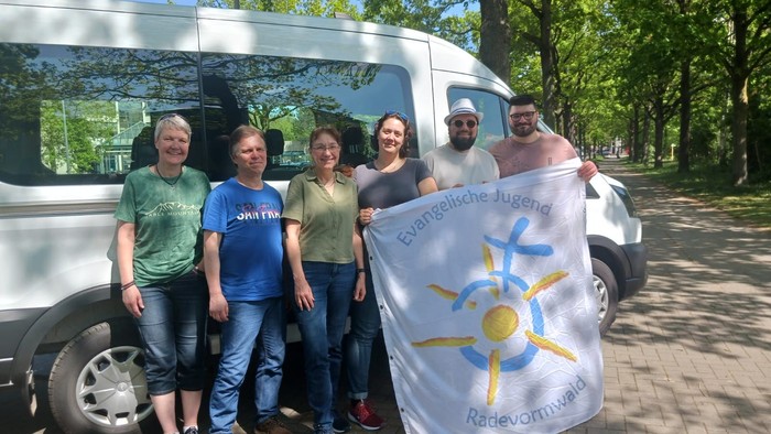 Eine Gruppe von Männern und Frauen mit einem Banner mit der Aufschrift: Evangelische Jugend Radevormwald vor einem Kleinbus auf einer Straße unter Bäumen.