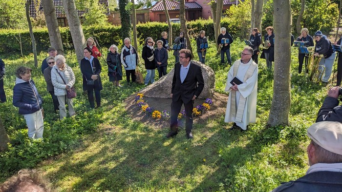 Eine Gruppe von Menschen versammelt sich in einem Garten, einige stehen, andere knien, ein Mann in weißem Gewand hält ein Buch.