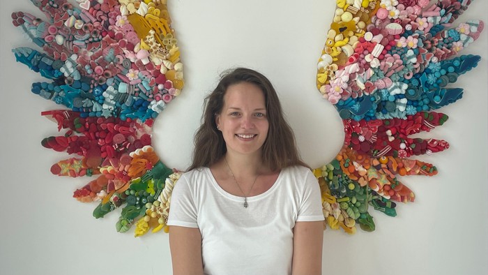 Eine Frau steht vor bunten Engelsflügeln an einer Wand.