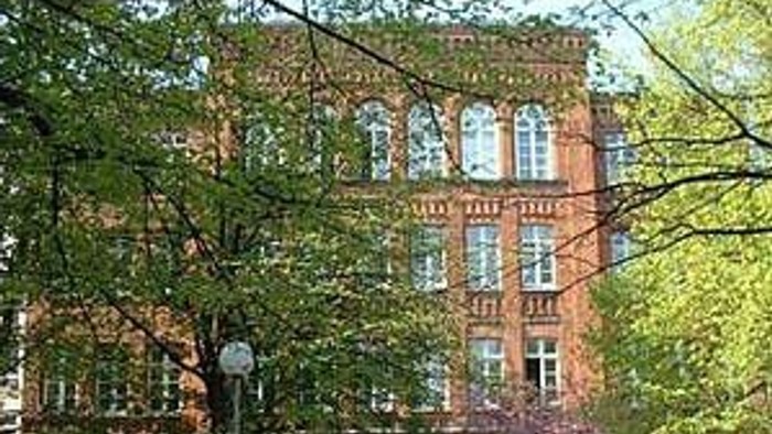 Das Bild zeigt ein altes Backsteingebäude mit grünen Fensterläden, umgeben von Bäumen und einem Laternenpfahl.