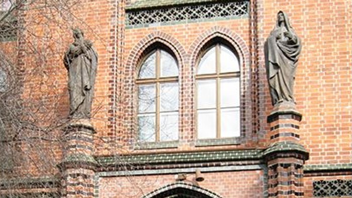 Das Bild zeigt den Eingang eines Ziegelgebäudes mit zwei Statuen und einem gewölbten Tor.