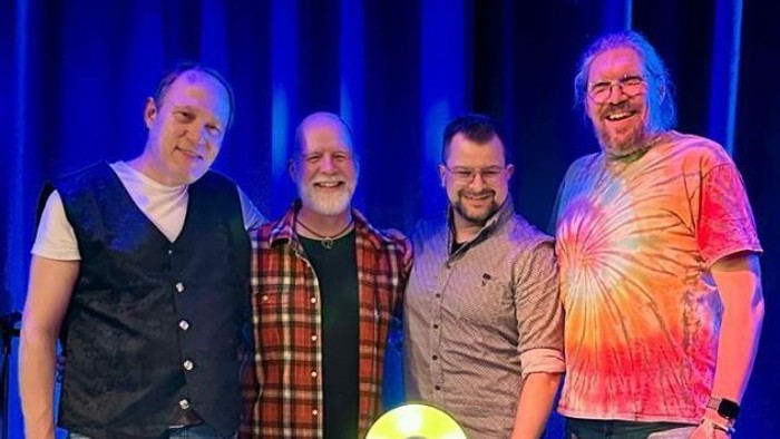 Vier Männer posieren gemeinsam auf der Bühne vor einem beleuchteten Ankerschild.