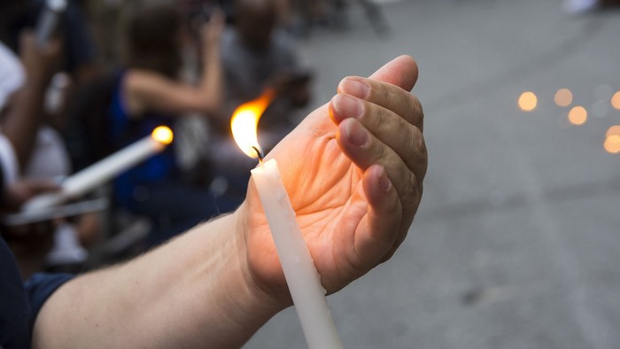 Eine Hand hält eine brennende Kerze, im Hintergrund sind weitere Menschen mit Kerzen zu sehen.