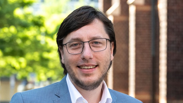 Ein Mann mit Brille und Anzug lächelt vor einem Gebäude.