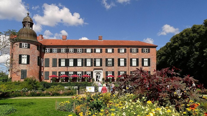 Ein großes, rotes Backsteingebäude mit grünen Fensterläden und Turm, umgeben von einem blühenden Garten mit bunten Blumen.