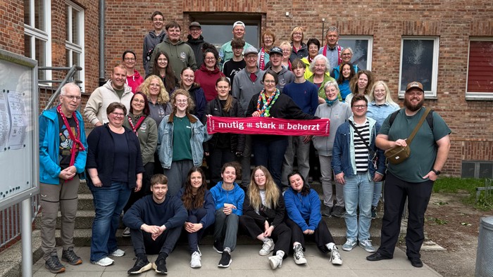 Gruppe von Personen, die vor einem Ziegelgebäude posieren, einige halten ein Schild.