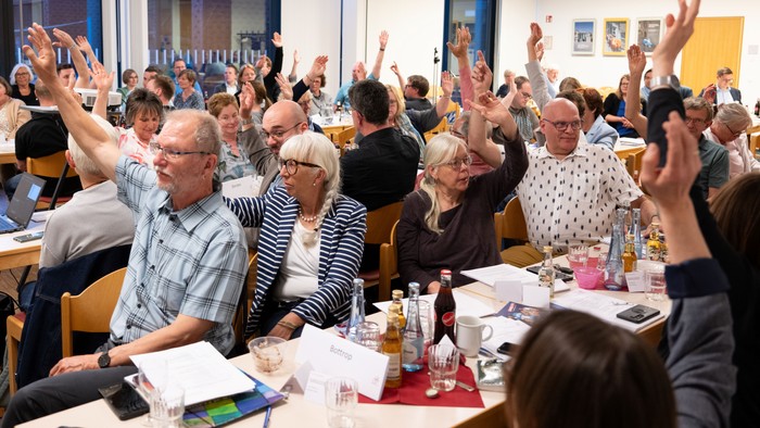 Eine Gruppe von Menschen sitzt an Tischen und hebt die Hände, offenbar bei einer Abstimmung oder Fragerunde.