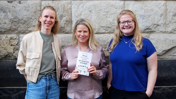 Drei Frauen stehen vor einer Steinmauer, eine hält ein Schild mit Text