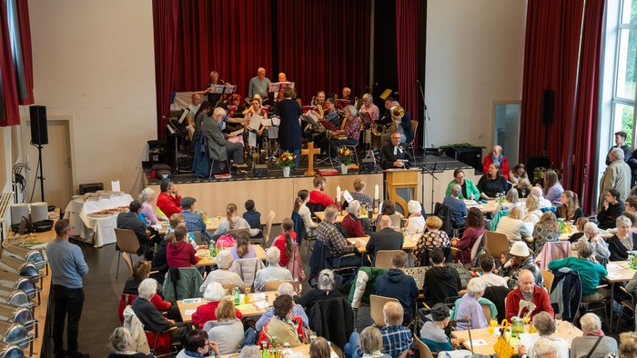 Eine große Gruppe von Menschen sitzt an Tischen und hört einem Orchester zu, das auf einer Bühne spielt.