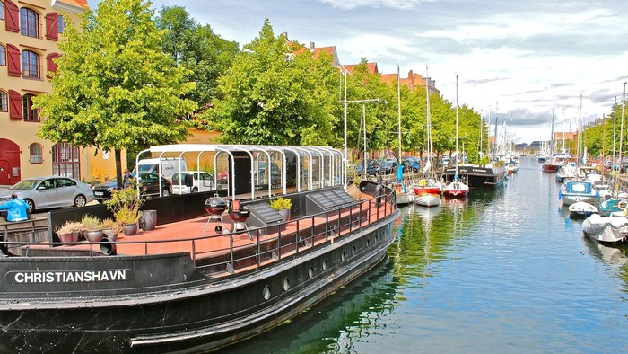 En stor båd med navnet Christianshavn ligger ved kaj i en kanal med andre både og farverige bygninger i baggrunden.