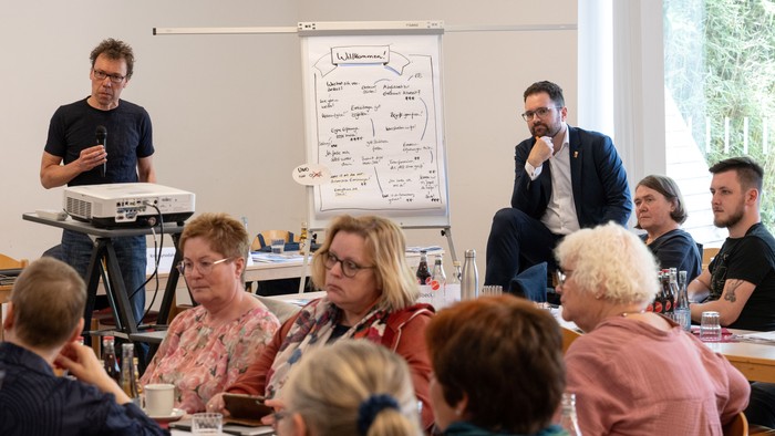 Ein Mann präsentiert vor einem Publikum an Tischen, während ein anderer Mann an einem Flipchart steht.