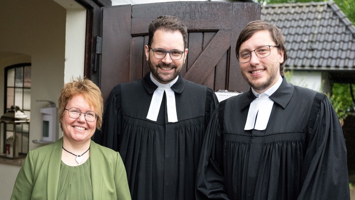 Drei Personen in formeller Kleidung stehen vor einem Haus