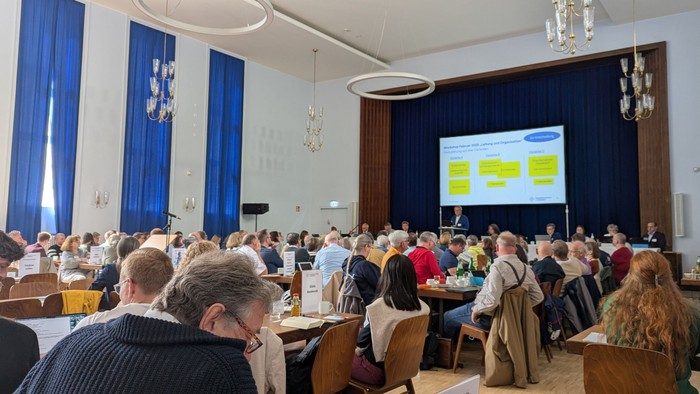 Eine große Gruppe von Menschen sitzt in einem Konferenzraum und schaut auf einen Bildschirm.