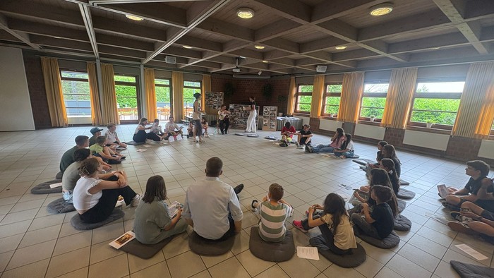 Eine Gruppe von Menschen sitzt im Kreis auf dem Boden in einem Raum mit großen Fenstern.