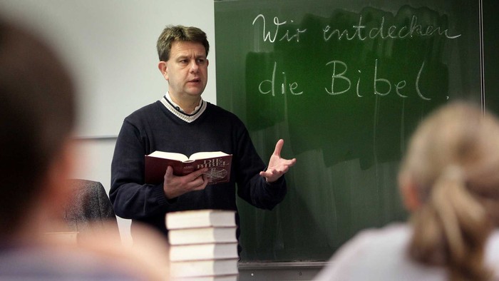 Mann hält Buch und referiert vor Gruppe vor grünem Brett mit Bibeltext