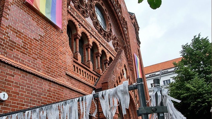 Passionskirche mit Regenbogenbanner. Davor Bauzaun mit schmalen, beschriebenen Zetteln.