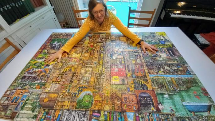 Eine Person posiert neben einem fertiggestellten großen Puzzle, das eine lebendige, detailreiche Stadtlandschaft zeigt.