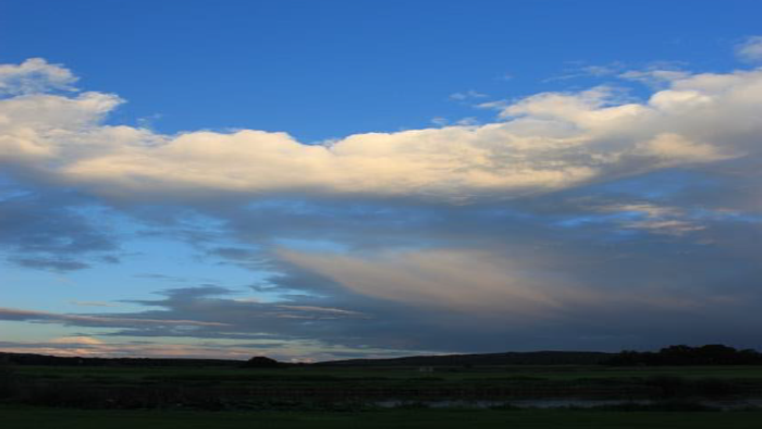 Eine ruhige Landschaft mit weitem Himmel, geprägt von dramatischen, zarten Wolken über offenen Feldern.