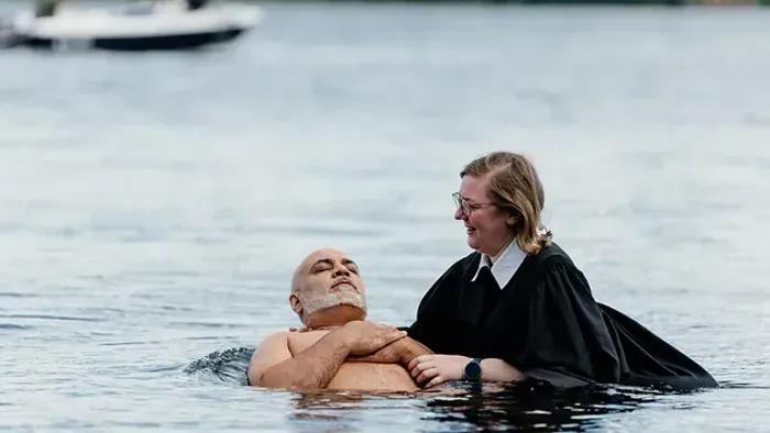 Zwei Personen im Wasser, eine in formeller Robe, die einem älteren Mann sanft beim Treiben hilft.