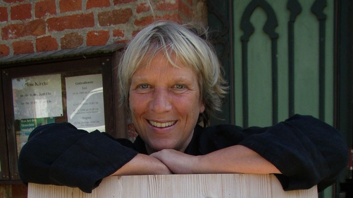 Eine Frau mit blondem Haar lehnt an einem Fenster und lächelt.