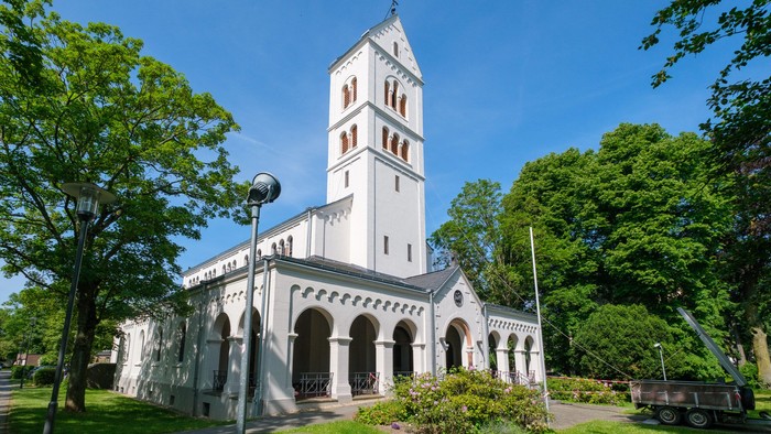 Weiße Kirche mit hohem Turm und Bogenfenstern, umgeben von Bäumen und Gras.
