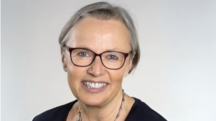 Eine Frau mit grauen Haaren und Brille lächelt in die Kamera.