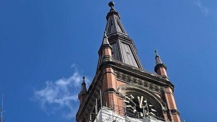 Ein hoher Kirchturm mit Uhr und spitzen Dachaufsätzen vor blauem Himmel