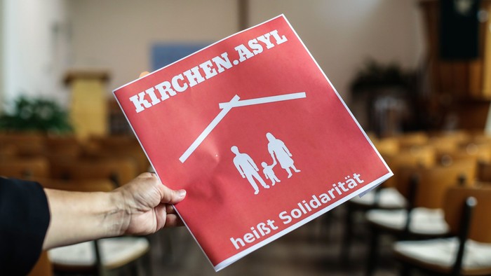 Person hält ein rotes Schild mit der Aufschrift "KIRCHEN.ASYL" und einer Familie mit einem Dach darüber