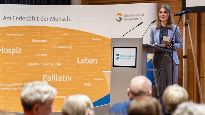 Eine Frau hält einen Vortrag vor einem Publikum, hinter ihr steht ein Banner mit dem Text "Am Ende zählt der Mensch."