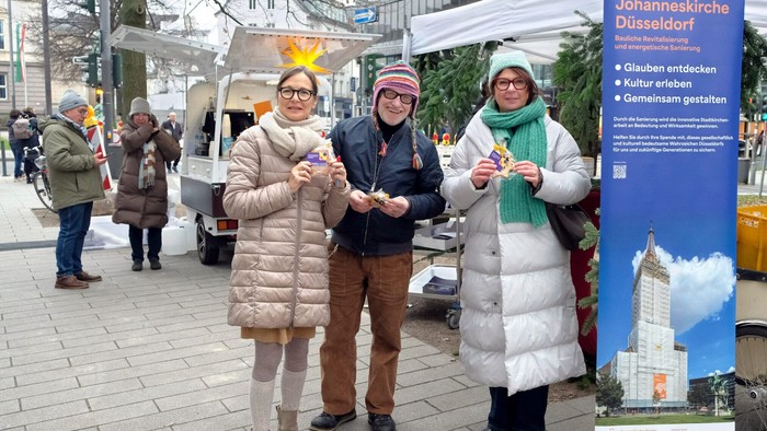 Drei Personen stehen vor einem Informationsstand der Johanneskirche Düsseldorf, jeder hält ein Getränk.