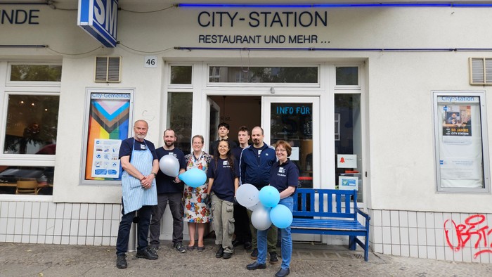 9 Mitarbeitende, das Team der City-Station, steht vor der Einrichtung. Einige von ihnen haben blaue Luftballons in der Hand, rechts steht eine blaue Bank vor der Glastür, 