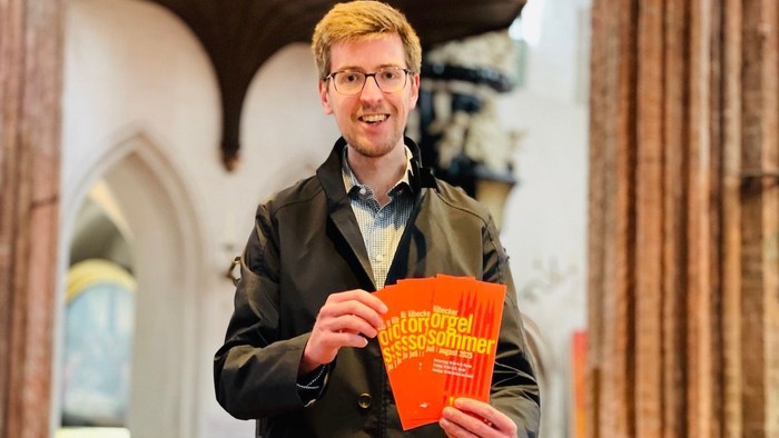 Christoph Arta, Leiter des Kirchenmusikbüros der Innenstadtkirchen, lädt zum 15. Lübecker Orgelsommer ein und hält Flyer mit dem Programm in den Händen. Er steht in der St.-Marien-Kirche in Lübeck.