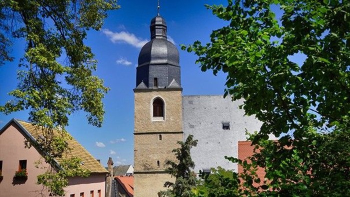Ein hoher Kirchturm mit schwarzem Dach zwischen üppigen grünen Bäumen und Häusern.