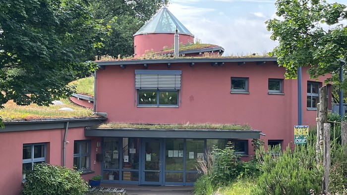 Ein rosa Gebäude mit grünem Dach und großen Fenstern, umgeben von Bäumen und Büschen.