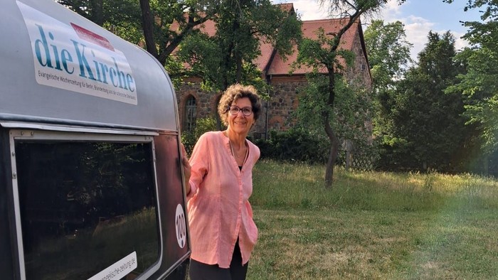Die Journalistin schaut hinter ihrem Campingwagen hervor. Im Hintergrund ist eine Kirche. 