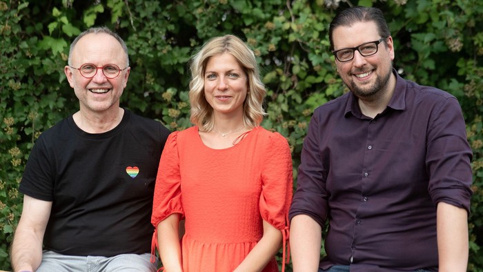 Drei Personen sitzen im Freien und lächeln in die Kamera