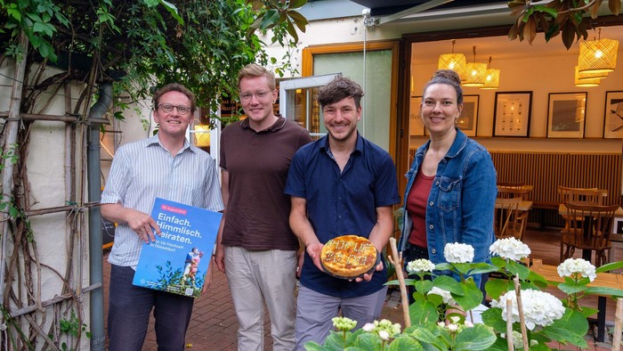 Vier Personen stehen vor einem Restaurant, zwei halten Bücher und einer eine Pizza.