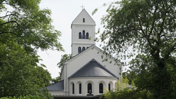 Weiße Kirche mit hohem Turm und Uhr, umgeben von Bäumen