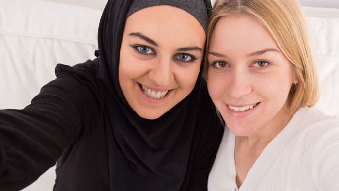 Selfiefoto von zwei lachenden Frauen, die Schulter an Schulter in die Kamera schauen. Die eine mit Kopftuch, die andere ohne.