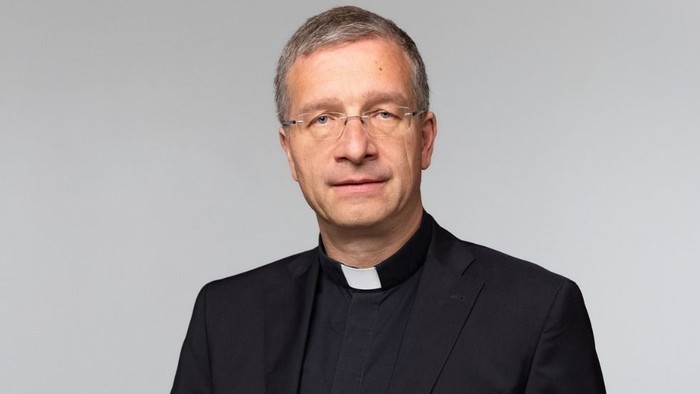 Ein Mann in schwarzer Priesterkleidung vor einem grauen Hintergrund.