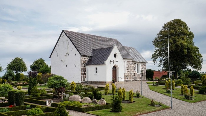 En lille, hvid kirke med sort tag og en smuk have med stene og planter.